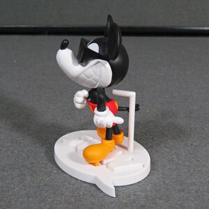 May include: A black, white, red, and yellow Mickey Mouse figurine with sunglasses. The figurine is standing on a white base with a speech bubble design. The figure is approximately 4 inches tall.