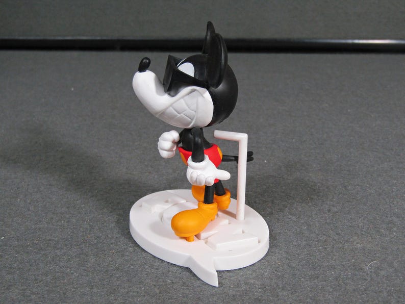 May include: A black, white, red, and yellow Mickey Mouse figurine with sunglasses. The figurine is standing on a white base with a speech bubble design. The figure is approximately 4 inches tall.