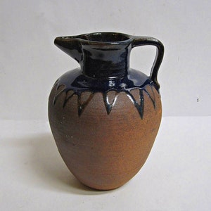 Puede incluir: Una jarra de cerámica marrón con un esmalte azul oscuro y un borde decorativo. La jarra tiene un cuerpo redondeado y un pequeño pico.