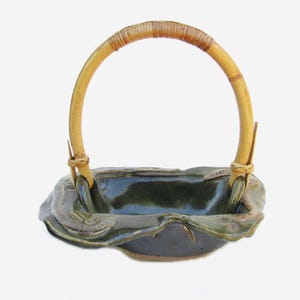 May include: A ceramic basket with a dark green glaze and a bamboo handle. The basket has an organic shape with a textured rim. The handle is made of natural bamboo. Suitable for decorative use.