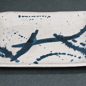 May include: Rectangular ceramic serving dish with a white base and dark blue abstract paint strokes and splatters. The dish has rounded corners and a slightly raised edge. A decorative piece for serving food.
