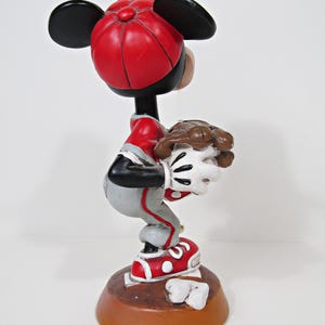 May include: A figurine of a cartoon character in a baseball uniform, wearing a red cap and shoes. The figure is in a crouched position, holding a baseball. The base is brown, and the figure has black ears.