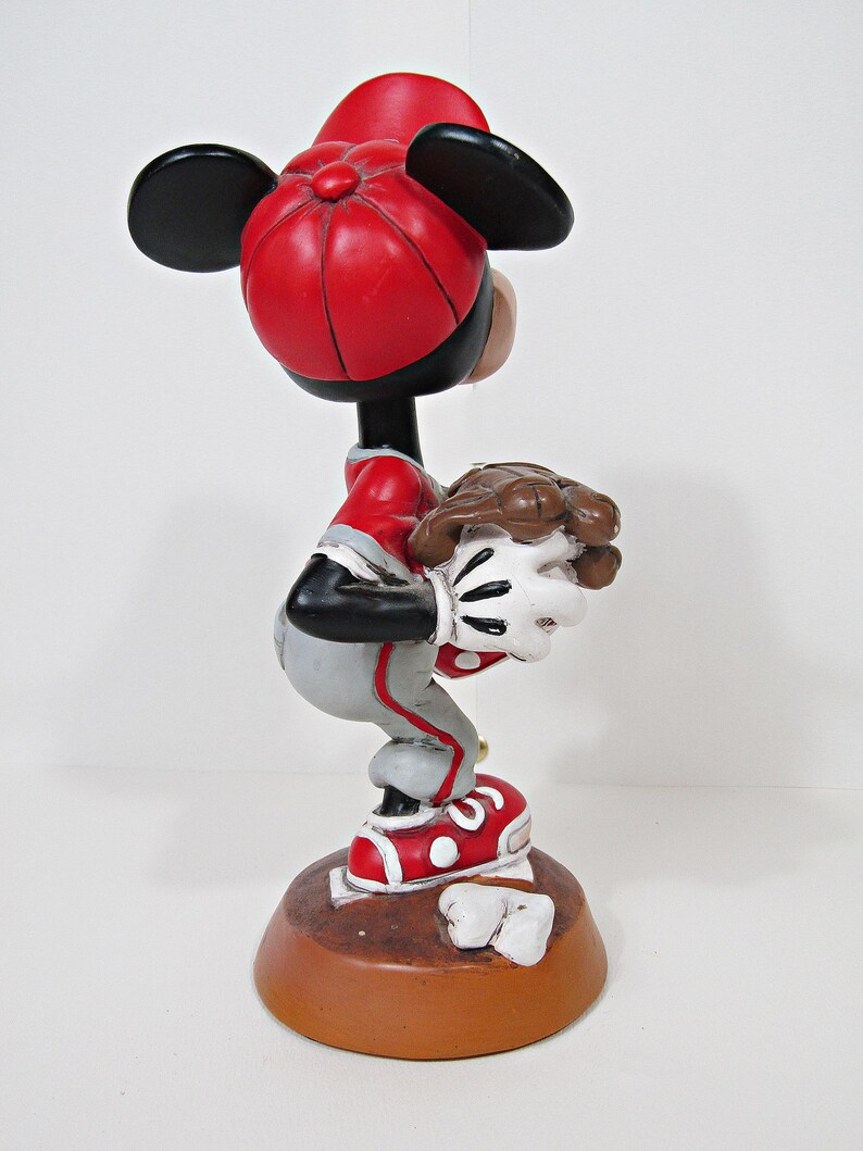 May include: A figurine of a cartoon character in a baseball uniform, wearing a red cap and shoes. The figure is in a crouched position, holding a baseball. The base is brown, and the figure has black ears.