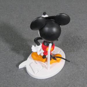 May include: A Mickey Mouse figurine with a black head and ears, white gloves, red shorts, and yellow shoes. The figure is standing on a white base with a stylized design. The figurine is a collectible item.