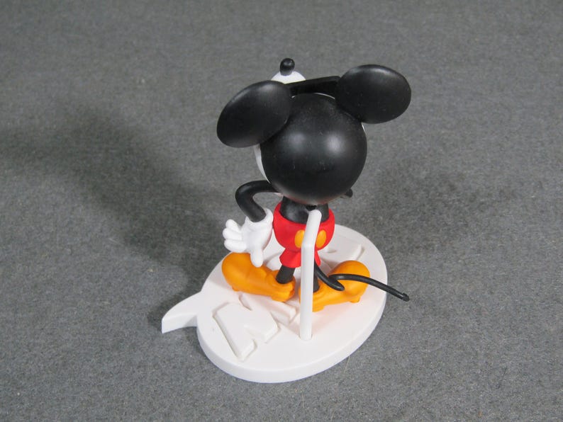 May include: A Mickey Mouse figurine with a black head and ears, white gloves, red shorts, and yellow shoes. The figure is standing on a white base with a stylized design. The figurine is a collectible item.