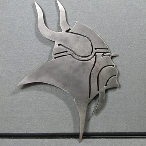 May include: A flat, silver-colored metal cutout depicting the profile of a Viking warrior wearing a horned helmet. The design features clean, laser-cut lines that define the facial features, beard, and helmet shape, creating a stylized silhouette.