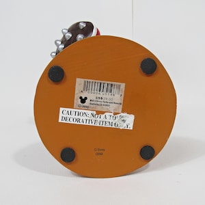 May include: A decorative item with an orange, circular base and four black rubber feet. The item has a sticker with the text "CAUTION: NOT A TOY DECORATIVE ITEM ONLY." and the Disney logo. The top portion of the item is visible, featuring a brown and silver design.