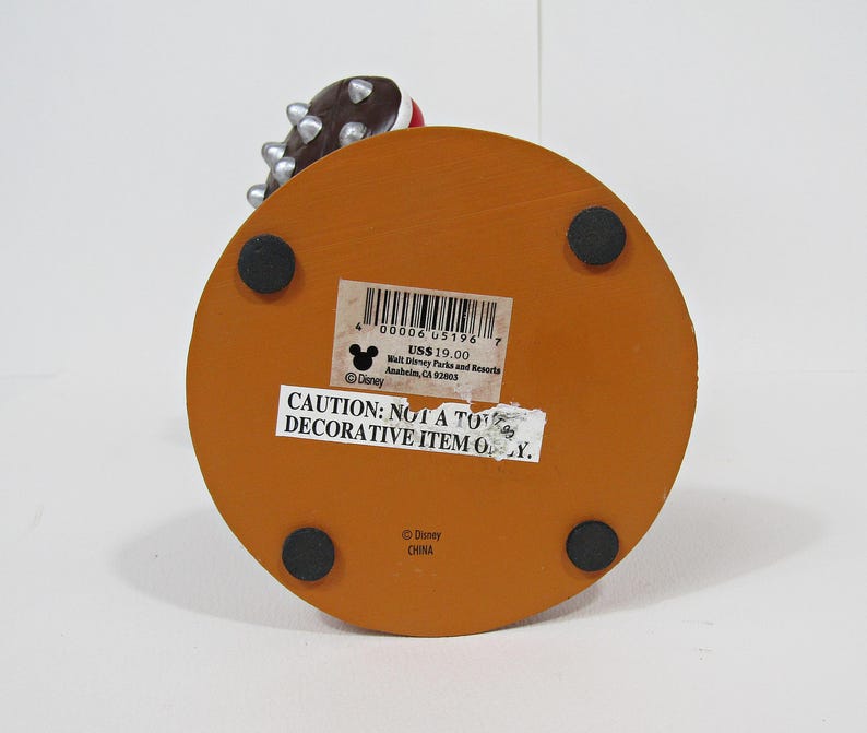 May include: A decorative item with an orange, circular base and four black rubber feet. The item has a sticker with the text "CAUTION: NOT A TOY DECORATIVE ITEM ONLY." and the Disney logo. The top portion of the item is visible, featuring a brown and silver design.