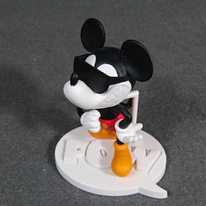 May include: A black, white, red, and yellow Mickey Mouse figurine wearing sunglasses. The figurine is standing on a white base with the word "POW" in raised letters. The figurine is approximately 4 inches tall.