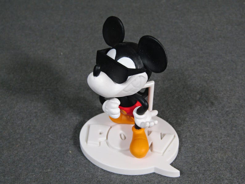 May include: A black, white, red, and yellow Mickey Mouse figurine wearing sunglasses. The figurine is standing on a white base with the word "POW" in raised letters. The figurine is approximately 4 inches tall.