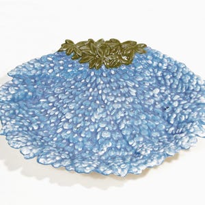 May include: A decorative blue and white ceramic dish with a textured surface. The dish has a scalloped edge and a cluster of green leaves at the top. The dish is approximately 10 inches in diameter.