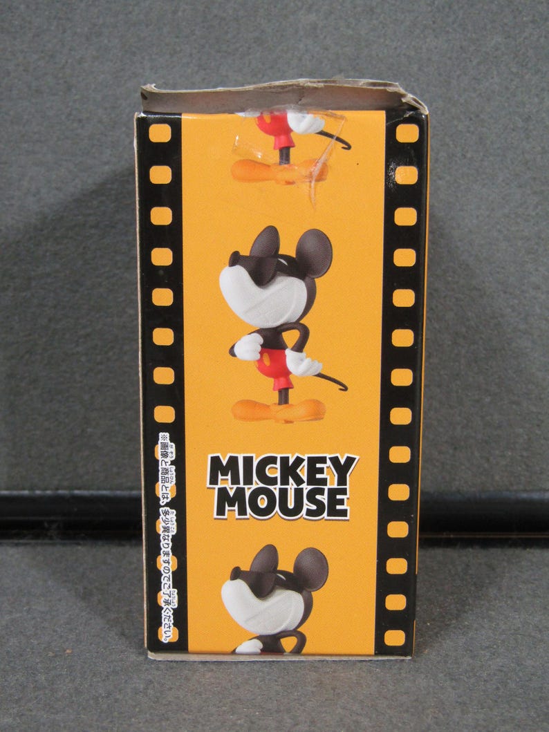 May include: A rectangular box with a yellow background featuring a cartoon image of Mickey Mouse. The box has a film strip design on the sides and the words "MICKEY MOUSE" in bold white letters.