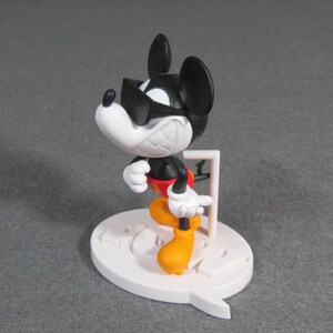 May include: A figurine of Mickey Mouse in a dynamic pose, wearing sunglasses. The figure has a black body, white face and hands, red shorts, and yellow shoes. It stands on a white base with a speech bubble design.