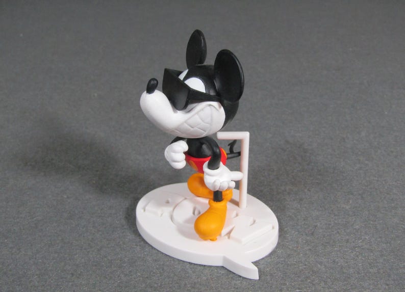 May include: A figurine of Mickey Mouse in a dynamic pose, wearing sunglasses. The figure has a black body, white face and hands, red shorts, and yellow shoes. It stands on a white base with a speech bubble design.