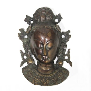 May include: A bronze-coloured metal sculpture of a woman's head with intricate details. The sculpture has a closed-eye expression and is adorned with floral and leaf-like designs.