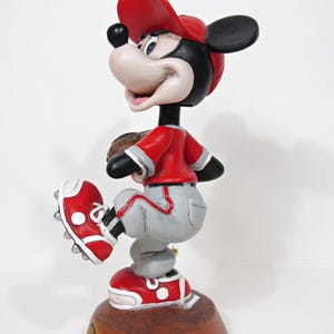 May include: A figurine of Mickey Mouse in a baseball uniform, wearing a red cap, jersey, and shoes, with grey trousers. The figurine is posed as if pitching a baseball. The base is brown and has the Disneyland Resort logo.