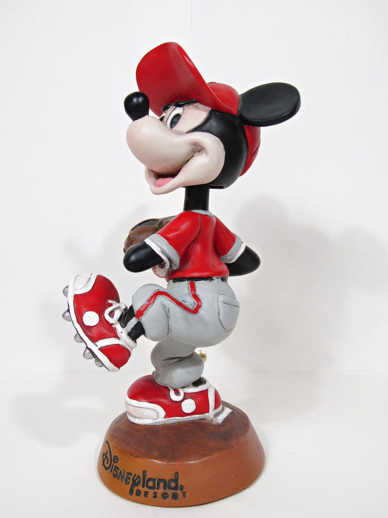 May include: A figurine of Mickey Mouse in a baseball uniform, wearing a red cap, jersey, and shoes, with grey trousers. The figurine is posed as if pitching a baseball. The base is brown and has the Disneyland Resort logo.