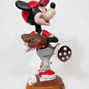 May include: A Mickey Mouse baseball figurine wearing a red cap and jersey, grey trousers, and red shoes. The figurine holds a baseball glove and ball. The brown base features the Disney logo.