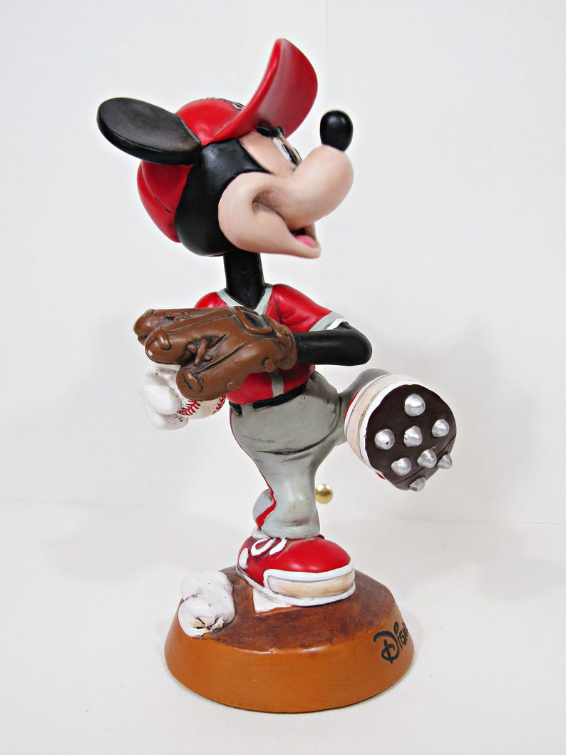 May include: A Mickey Mouse baseball figurine wearing a red cap and jersey, grey trousers, and red shoes. The figurine holds a baseball glove and ball. The brown base features the Disney logo.