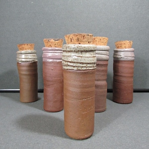 May include: A collection of six handmade ceramic bottles with cork stoppers. The bottles are cylindrical, with a reddish-brown base and a lighter, textured top. The corks are a natural tan color.