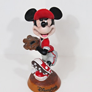 May include: A Mickey Mouse figurine in a baseball uniform, holding a glove and wearing a red cap and shoes. The figurine is on a brown base with "Disneyland Resort" printed on it. The figure is approximately 15 cm tall.