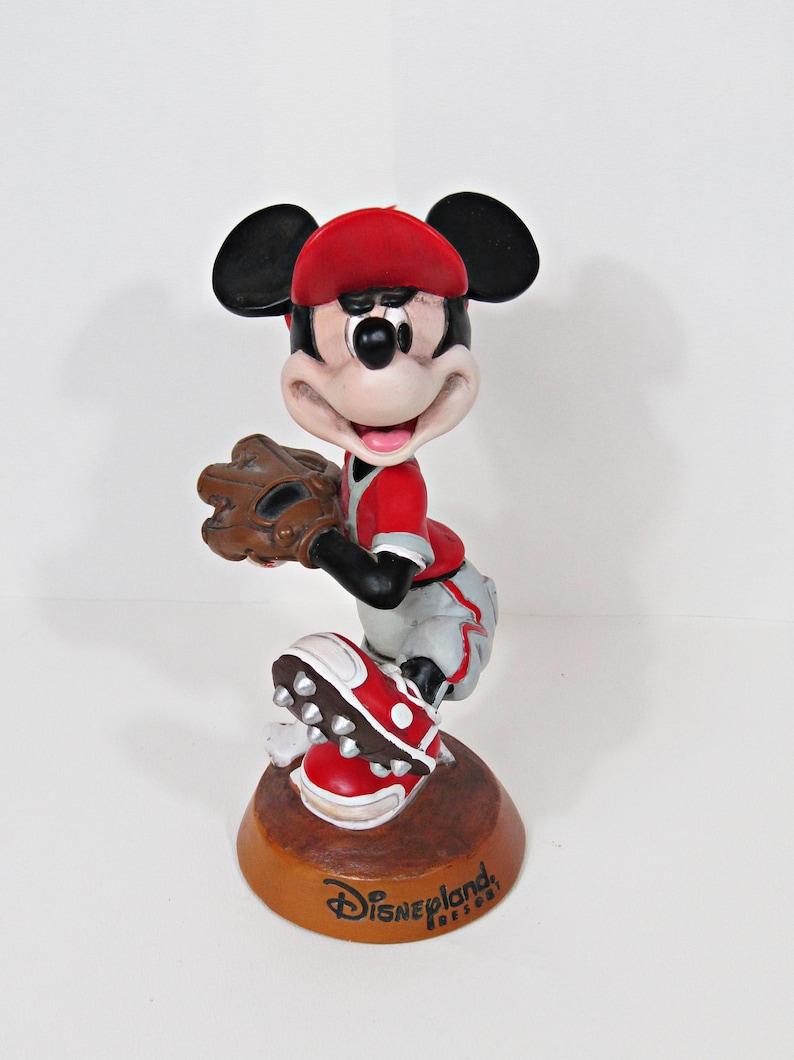 May include: A Mickey Mouse figurine in a baseball uniform, holding a glove and wearing a red cap and shoes. The figurine is on a brown base with "Disneyland Resort" printed on it. The figure is approximately 15 cm tall.