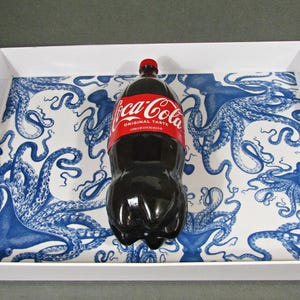 May include: A white serving tray with a blue octopus design holds a Coca-Cola bottle. The bottle has a red label with white text. The tray has handles and a rectangular shape.