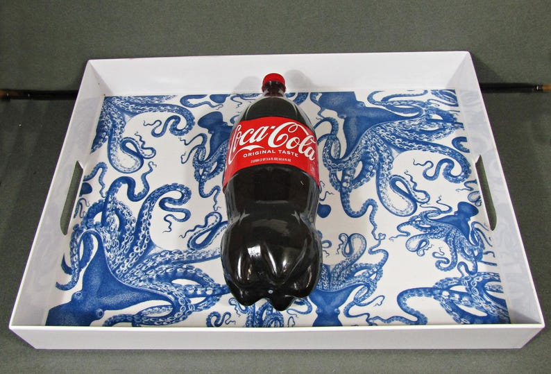 May include: A white serving tray with a blue octopus design holds a Coca-Cola bottle. The bottle has a red label with white text. The tray has handles and a rectangular shape.