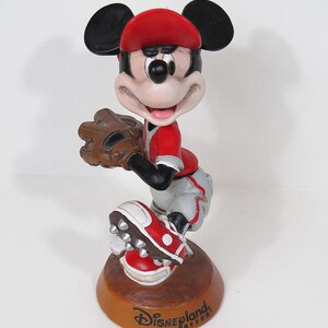 May include: A Mickey Mouse figurine in a baseball uniform, holding a glove. The figure wears a red cap, jersey, and shoes, with grey trousers. The base is brown with "Disneyland Resort" text.