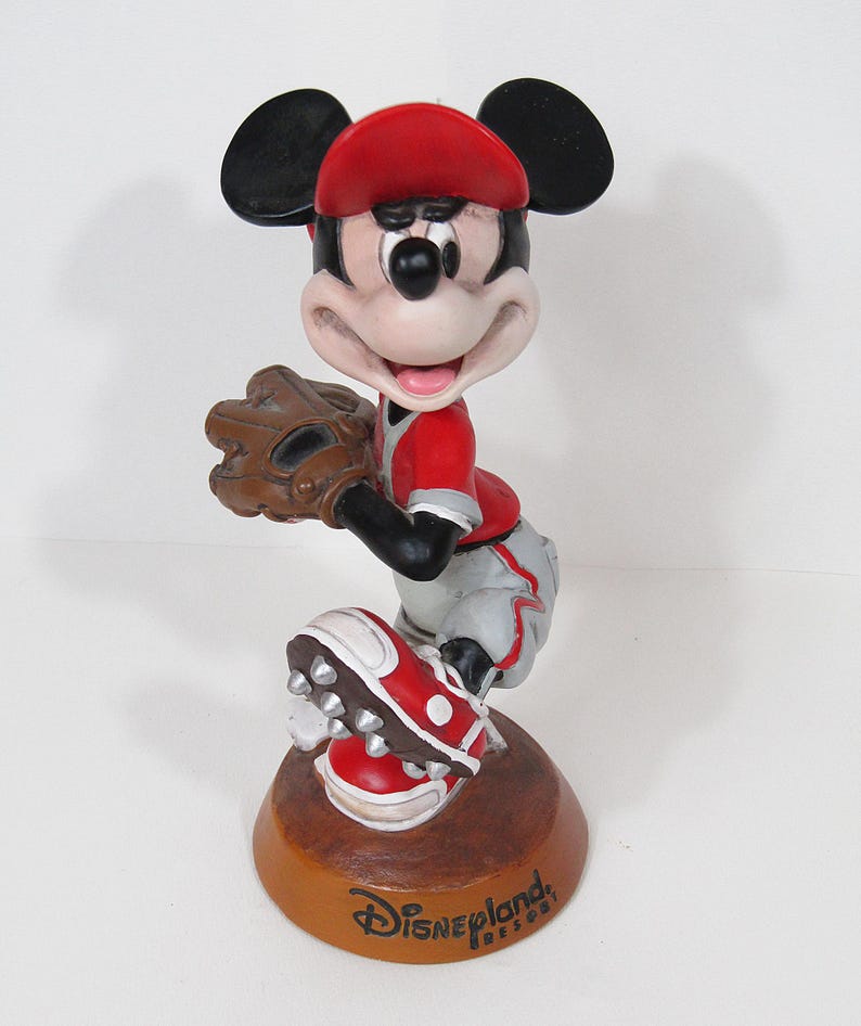 May include: A Mickey Mouse figurine in a baseball uniform, holding a glove. The figure wears a red cap, jersey, and shoes, with grey trousers. The base is brown with "Disneyland Resort" text.