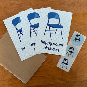 May include: Greeting cards with a blue folding chair design and the text "happy sober birthday." The cards have a light blue polka dot background. Also included are matching stickers and a brown envelope.