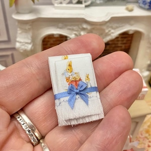 Può includere: Un set in miniatura di due asciugamani bianchi con bordino blu e un fiocco blu. Gli asciugamani presentano un'immagine stampata di Peter Rabbit e dei suoi amici.