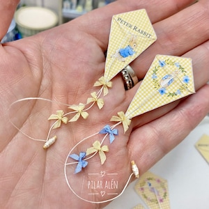 May include: Two miniature kites featuring yellow gingham patterns and Peter Rabbit illustrations. Each kite is decorated with small yellow and blue bows along the string. The kites are held in a hand.