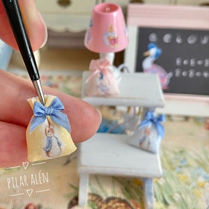 K&ouml;nnte beinhalten: Ein Set aus drei Miniatur-Stofftaschen mit blauen Schleifen. Die Taschen sind gelb mit einem wei&szlig;en Hasendesign. Die Taschen befinden sich auf einem wei&szlig;en Holzregal mit einer rosa Lampe und einer Tafel.