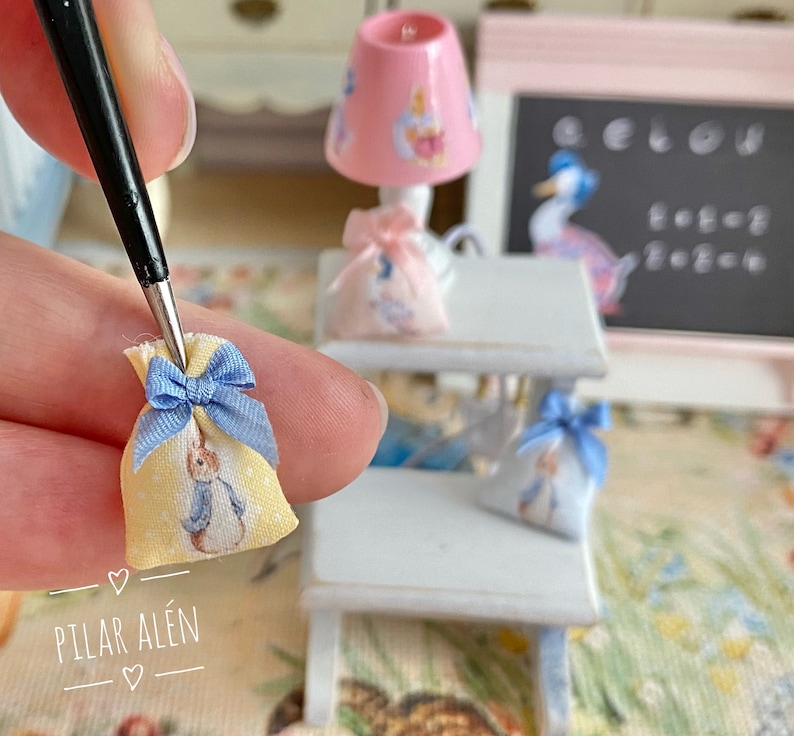 K&ouml;nnte beinhalten: Ein Set aus drei Miniatur-Stofftaschen mit blauen Schleifen. Die Taschen sind gelb mit einem wei&szlig;en Hasendesign. Die Taschen befinden sich auf einem wei&szlig;en Holzregal mit einer rosa Lampe und einer Tafel.