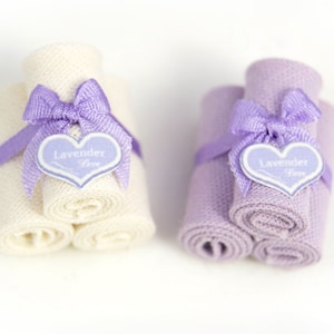 May include: Two sets of rolled towels, one set is white and the other is purple. Both sets are tied with a purple ribbon and have a heart-shaped tag that says "Lavender Love".
