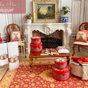 May include: Miniature room setting with a fireplace, chairs, and a rug. Red plaid gift boxes with red bows are stacked. White towels with a deer design hang on a rack. Pillows with deer designs are on the chairs. Books and a decorative painting are also present.