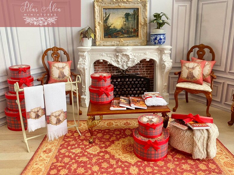 May include: Miniature room setting with a fireplace, chairs, and a rug. Red plaid gift boxes with red bows are stacked. White towels with a deer design hang on a rack. Pillows with deer designs are on the chairs. Books and a decorative painting are also present.