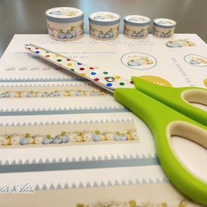 May include: A sheet of paper with a blue and white patterned border and a row of small pumpkins. The paper is labeled "Autumn by Pilar Alin" and includes instructions to print, cut, glue, and enjoy. A pair of green scissors with colorful polka dots is on top of the paper.