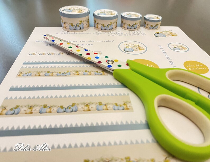 May include: A sheet of paper with a blue and white patterned border and a row of small pumpkins. The paper is labeled "Autumn by Pilar Alin" and includes instructions to print, cut, glue, and enjoy. A pair of green scissors with colorful polka dots is on top of the paper.