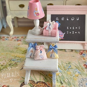 K&ouml;nnte beinhalten: Ein wei&szlig;er Holzhocker mit drei blauen und wei&szlig;en Stoffbeuteln, die an der Oberseite h&auml;ngen. Eine rosa Lampe mit wei&szlig;em Sockel und ein wei&szlig;er und rosa Stoffbeutel befinden sich auf der oberen Ablage. Eine rosa Kreidetafel mit den Buchstaben aeiou und den Gleichungen 1+1=2 und 2+2=4 befindet sich an der Wand hinter dem Hocker.