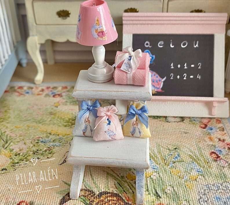 K&ouml;nnte beinhalten: Ein wei&szlig;er Holzhocker mit drei blauen und wei&szlig;en Stoffbeuteln, die an der Oberseite h&auml;ngen. Eine rosa Lampe mit wei&szlig;em Sockel und ein wei&szlig;er und rosa Stoffbeutel befinden sich auf der oberen Ablage. Eine rosa Kreidetafel mit den Buchstaben aeiou und den Gleichungen 1+1=2 und 2+2=4 befindet sich an der Wand hinter dem Hocker.