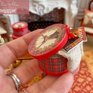 May include: Miniature red and plaid round box with a deer illustration on the lid. The box contains a small magazine and a white cloth. The box is held in a hand. Other miniature items are in the background.