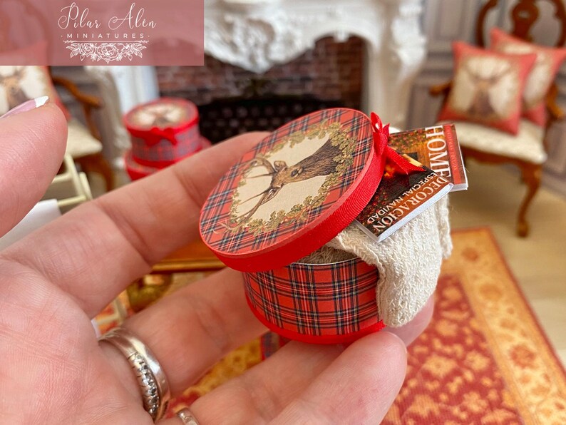 May include: Miniature red and plaid round box with a deer illustration on the lid. The box contains a small magazine and a white cloth. The box is held in a hand. Other miniature items are in the background.