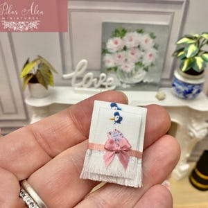 Può includere: Un asciugamano rettangolare in miniatura di colore bianco con bordo inferiore sfrangiato, caratterizzato da una piccola grafica di uccellino blu e un nastro rosa annodato a fiocco al centro. Il tessuto ha un motivo floreale blu e texture liscia.