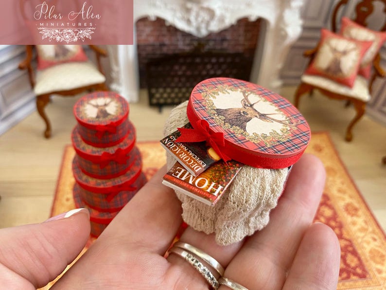 May include: Miniature Christmas-themed decorative boxes and accessories. The boxes are red with a plaid pattern and a deer illustration. A small book with the word "HOME" is visible. The scene includes miniature furniture and a fireplace.