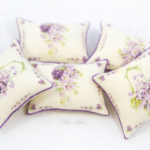 May include: Five decorative throw pillows with a floral pattern. The pillows are cream colored with a purple border and feature a watercolor design of purple flowers. The pillows are arranged in a pile on a white surface.