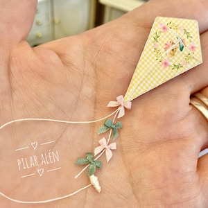 May include: A miniature kite with a yellow gingham pattern and floral accents, held in a hand. The kite is adorned with pink and green bows and a white string. The text "PILAR ALÉN" is visible.