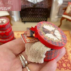 May include: Miniature red and plaid round box with a deer illustration on the lid, containing a beige knitted item and small books. A stack of similar boxes is visible in the background. The box is held in a hand.