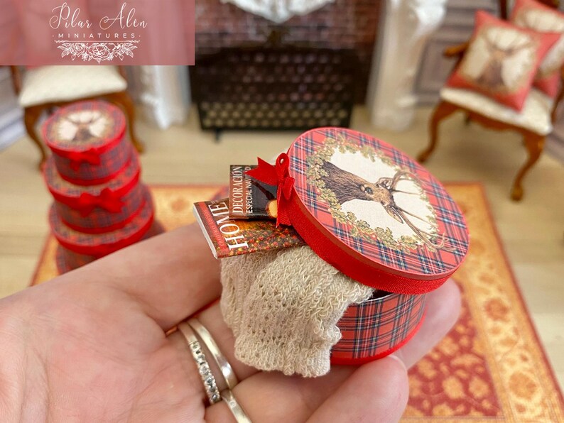 May include: Miniature red and plaid round box with a deer illustration on the lid, containing a beige knitted item and small books. A stack of similar boxes is visible in the background. The box is held in a hand.